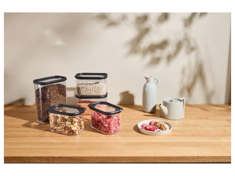 Transparente Lebensmittelbehälter mit Kaffeebohnen, Nüssen, Linsen und Trockenfrüchten auf einem Holztisch.