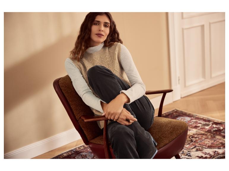 Frau in beiger Strickweste und dunklen Jeans sitzt in einem Sessel.