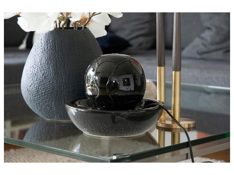 Schwarzer Tischbrunnen und Vase mit weißen Blumen auf einem Glastisch.