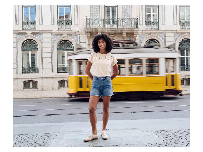 Frau in besticktem T-Shirt und Jeans-Shorts vor einer fahrenden gelben Straßenbahn.