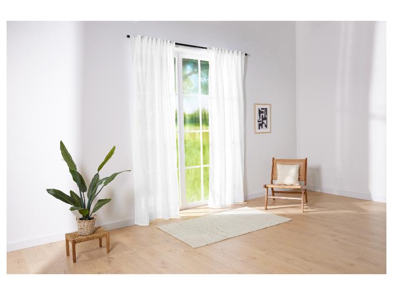 Lange weiße Vorhänge und ein Rattanstuhl in einem hellen Raum.