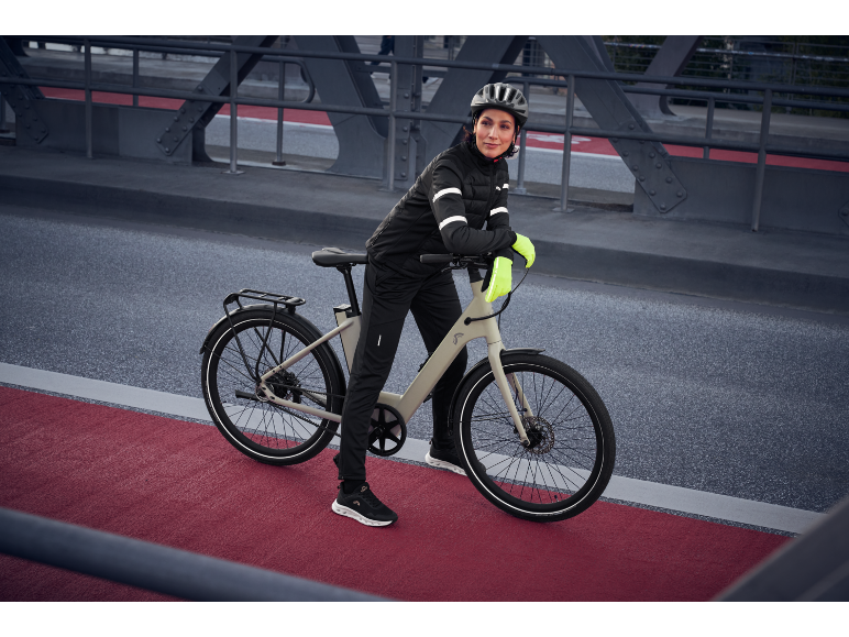 Frau in schwarzer Radsportbekleidung und gelben Handschuhen mit E-Bike auf einer Brücke.