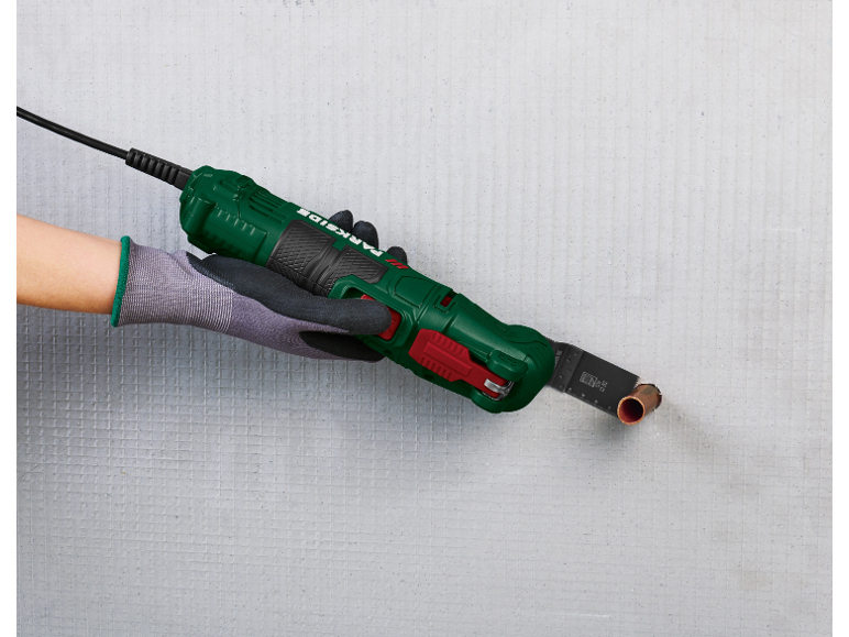 Eine Hand in einem Arbeitshandschuh hält ein Multifunktionswerkzeug, das ein Kupferrohr in einer Wand schneidet.