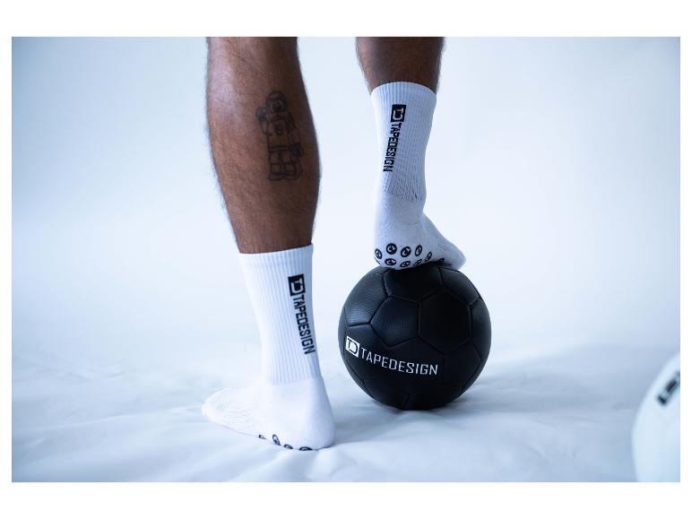 Person in weißen Sportsocken mit Anti-Rutsch-Noppen, Fuß auf schwarzem Fußball.