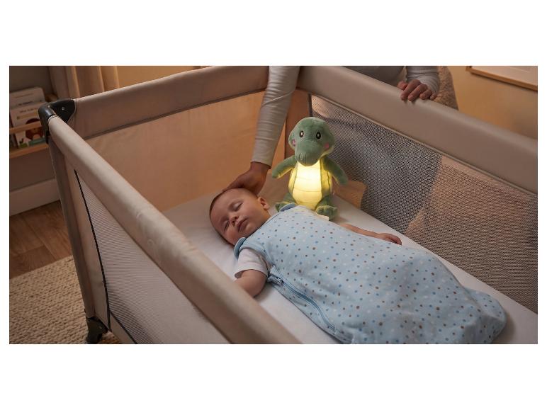 Baby schläft in einem Reisebett mit leuchtendem Krokodil-Nachtlicht, Elternhand in der Nähe.