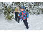 Zwei glückliche Kinder in bunten Schneeanzügen rennen im Schnee zwischen Bäumen.