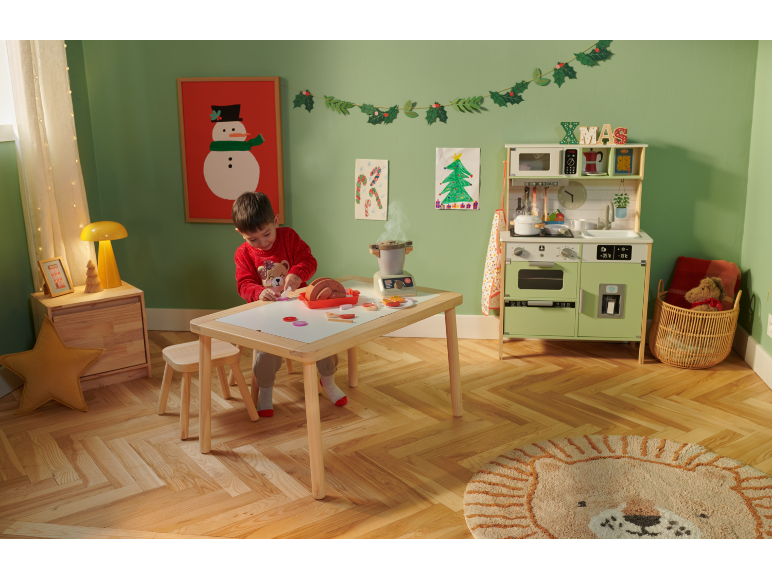 Ein Kind spielt mit einer Spielzeugküche und einer Sitzgruppe aus Holz in einem weihnachtlichen Zimmer.
