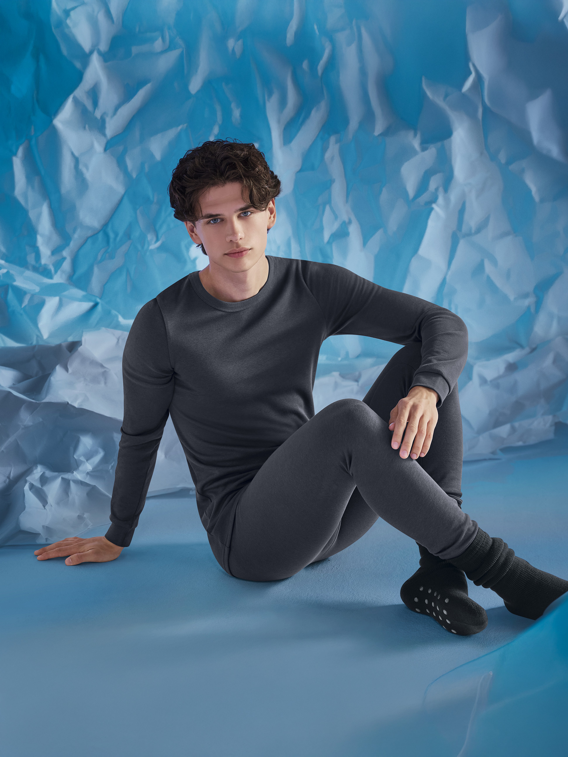 Mann in grauer Thermounterwäsche, sitzend vor einem zerknitterten blauen Hintergrund.