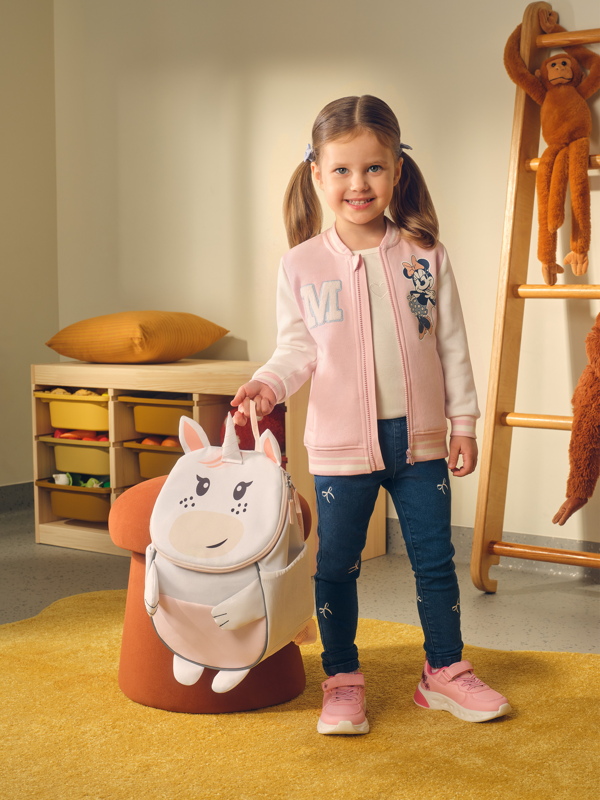 Lächelndes kleines Mädchen mit Einhorn-Rucksack, rosa Jacke und Jeans.