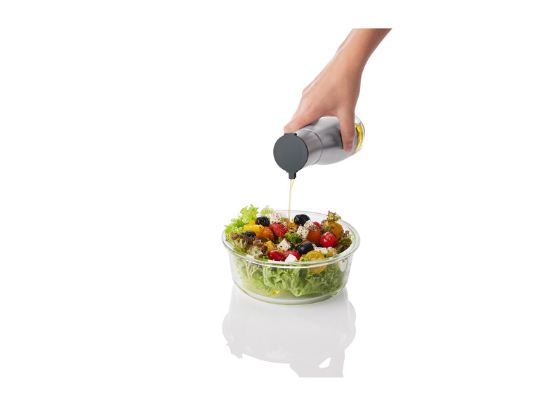 Eine Hand gießt Öl aus einer Flasche auf einen Salat in einer Glasschüssel.