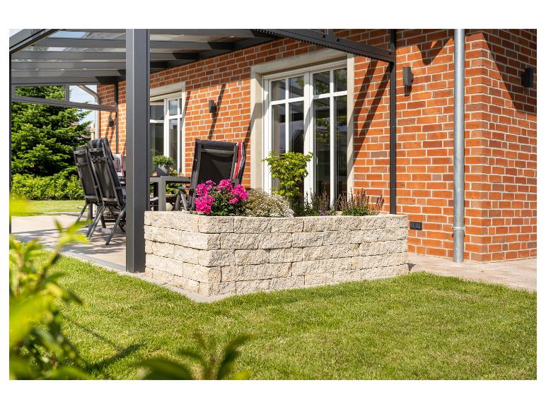Terrasse mit Glasüberdachung, Gartenmöbeln und Blumenbeet.