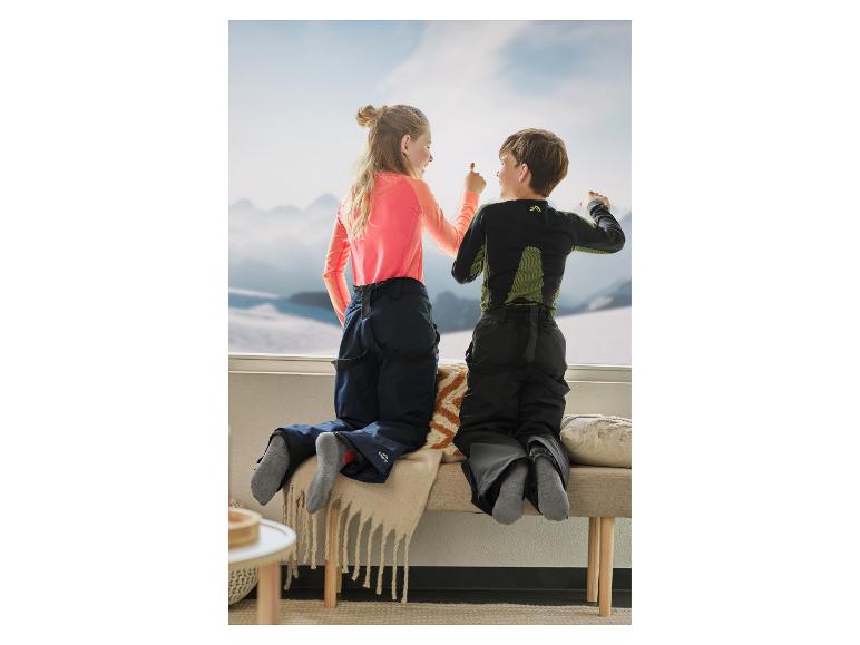 Zwei Kinder in Skibekleidung schauen aus einem Fenster auf verschneite Berge.