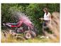 Lächelnde Frau reinigt rotes Quad mit Gartenschlauch in grünem Garten.