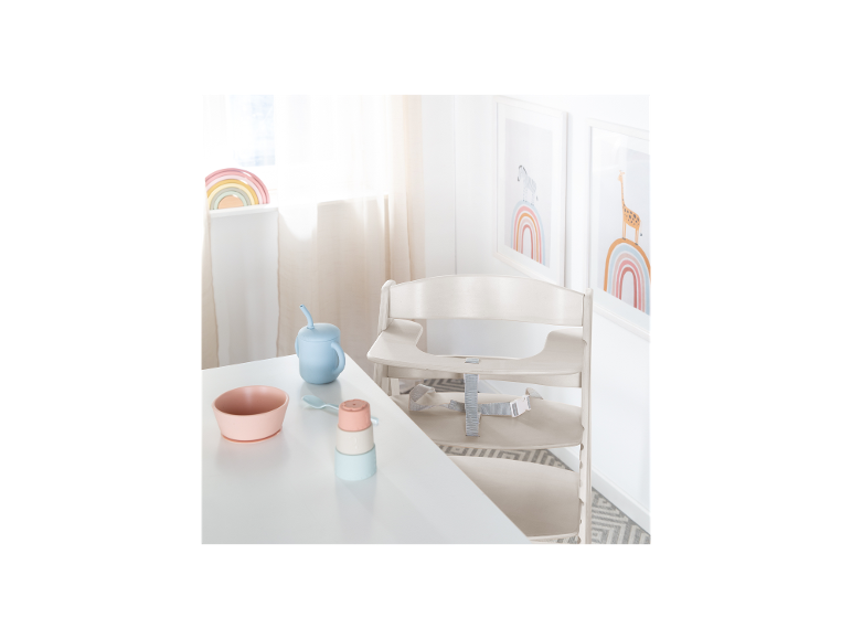 Ein Babyhochstuhl mit Tablett und Sicherheitsgurt, neben einem Tisch mit Babyfütterungsartikeln.