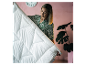 Frau hält eine weiße Bettdecke mit geometrischem Muster vor rosa Hintergrund und Pflanze.