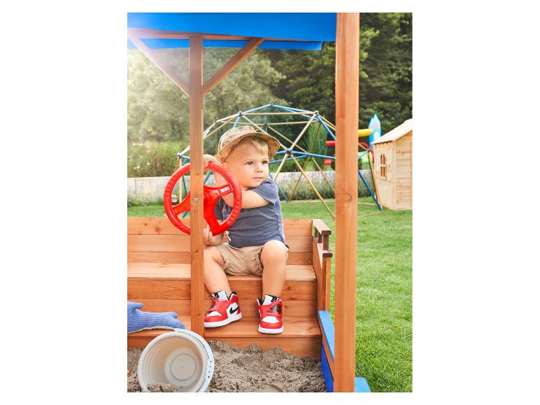 Kleinkind spielt in einem Holzsandkasten mit rotem Lenkrad, trägt Hut und Turnschuhe.