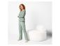 Frau in grünem Pyjama mit gemusterter Hose, stehend neben einem weißen Sessel.