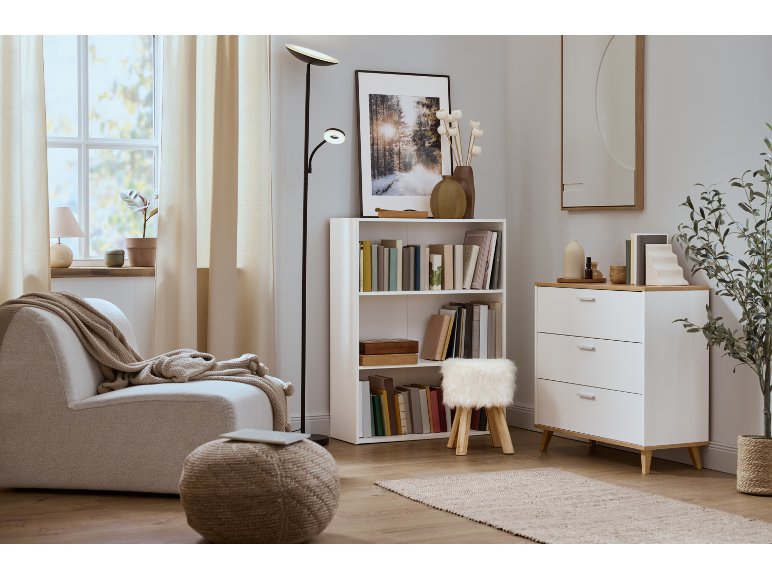 Ein helles Wohnzimmer mit beigem Sofa, weißem Bücherregal und Kommode mit Schubladen.