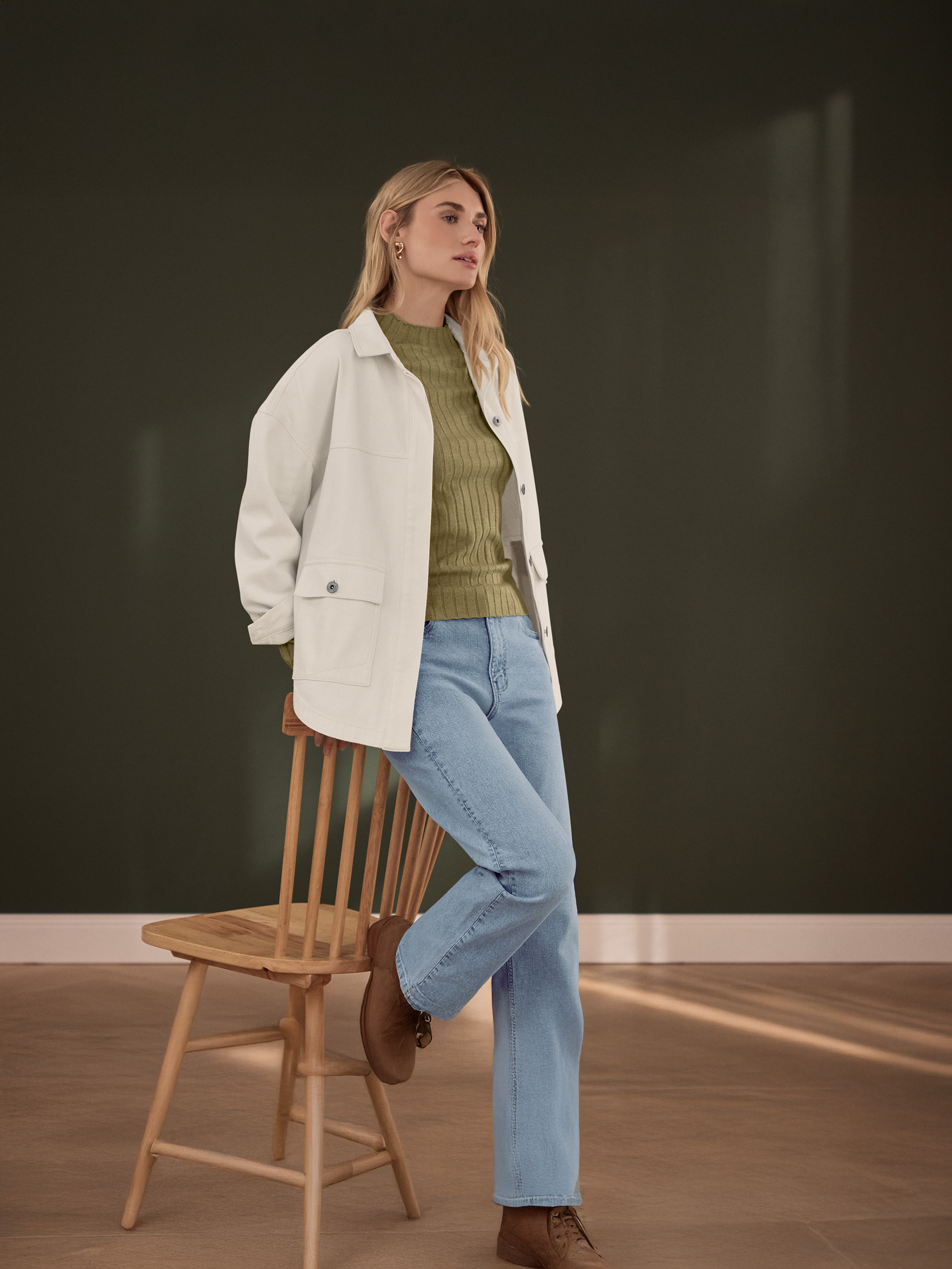 Frau in weißer Jacke, grünem Rippstricktop und hellblauen Jeans, lehnt an einem Holzstuhl.