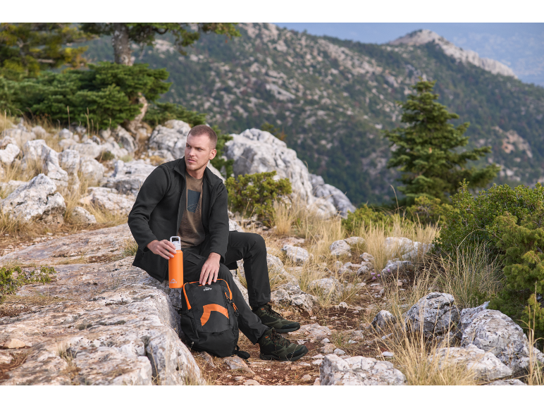 Mann in Fleecejacke und Trekkinghose sitzt auf einem Felsen mit Rucksack und Flasche.