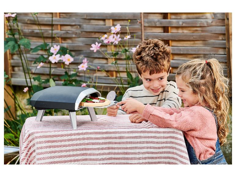 Zwei Kinder bereiten Spielzeugpizza in einem Outdoor-Ofen auf einem gestreiften Tischtuch zu.