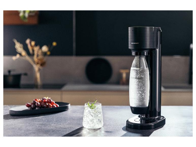 Ein schwarzer SodaStream Sprudler mit einer Flasche Sprudelwasser, Trauben und einem Glas Eisgetränk auf einer Küchenarbeitsplatte.