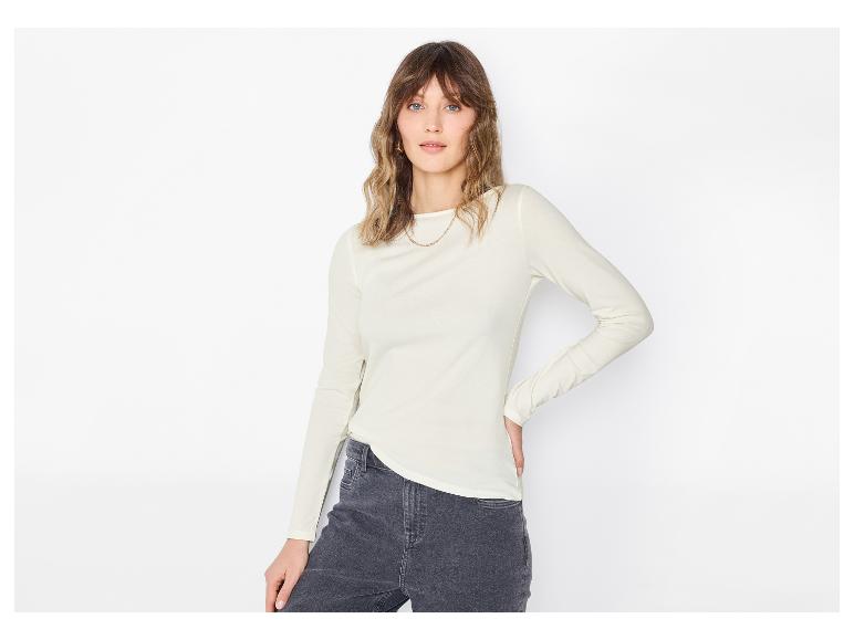 Frau in einer hellen Langarmbluse und dunklen Jeans, posiert vor weißem Hintergrund.
