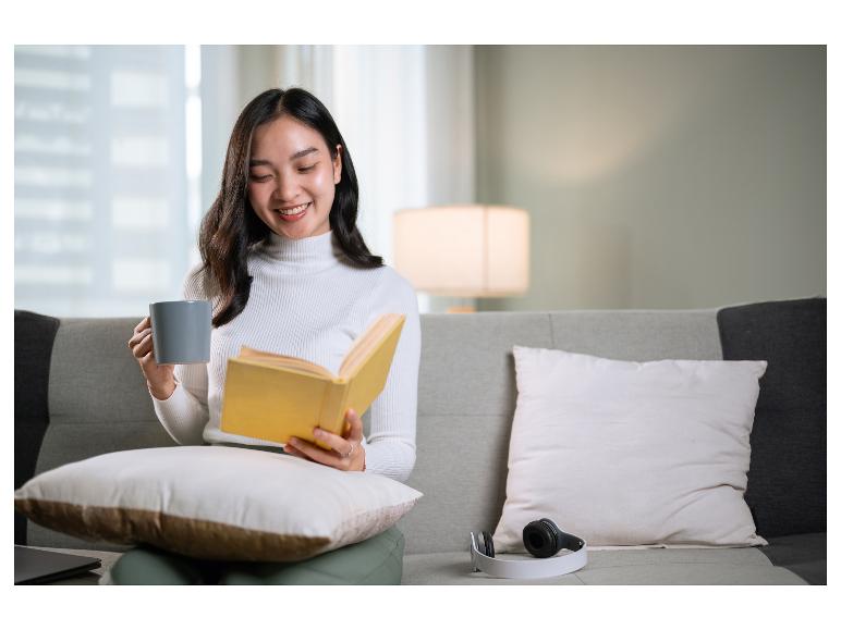 Lächelnde Frau liest ein Buch und hält eine Tasse auf einem Sofa.