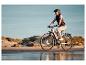 Frau auf E-Bike am Strand mit Dünen im Hintergrund.