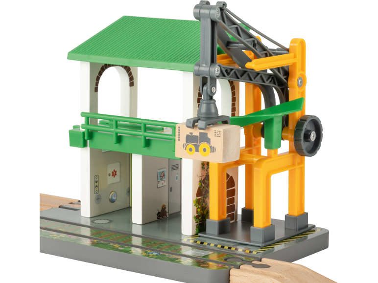 Hölzerner Spielzeug-Bahnhof mit grünem Dach und gelbem Kran, der eine Holzkiste hebt.
