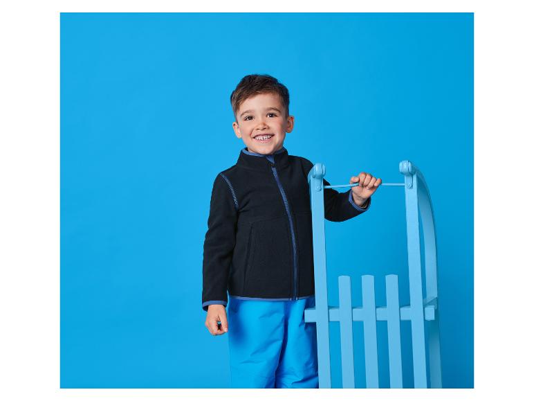 Lächelnder Junge in Jacke und Skihose, neben einem blauen Schlitten.
