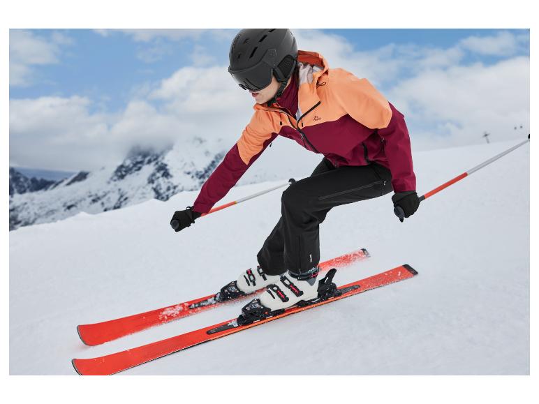 Eine Frau fährt Ski auf einem verschneiten Hang in einer orange-burgunderroten Jacke und schwarzen Hosen.