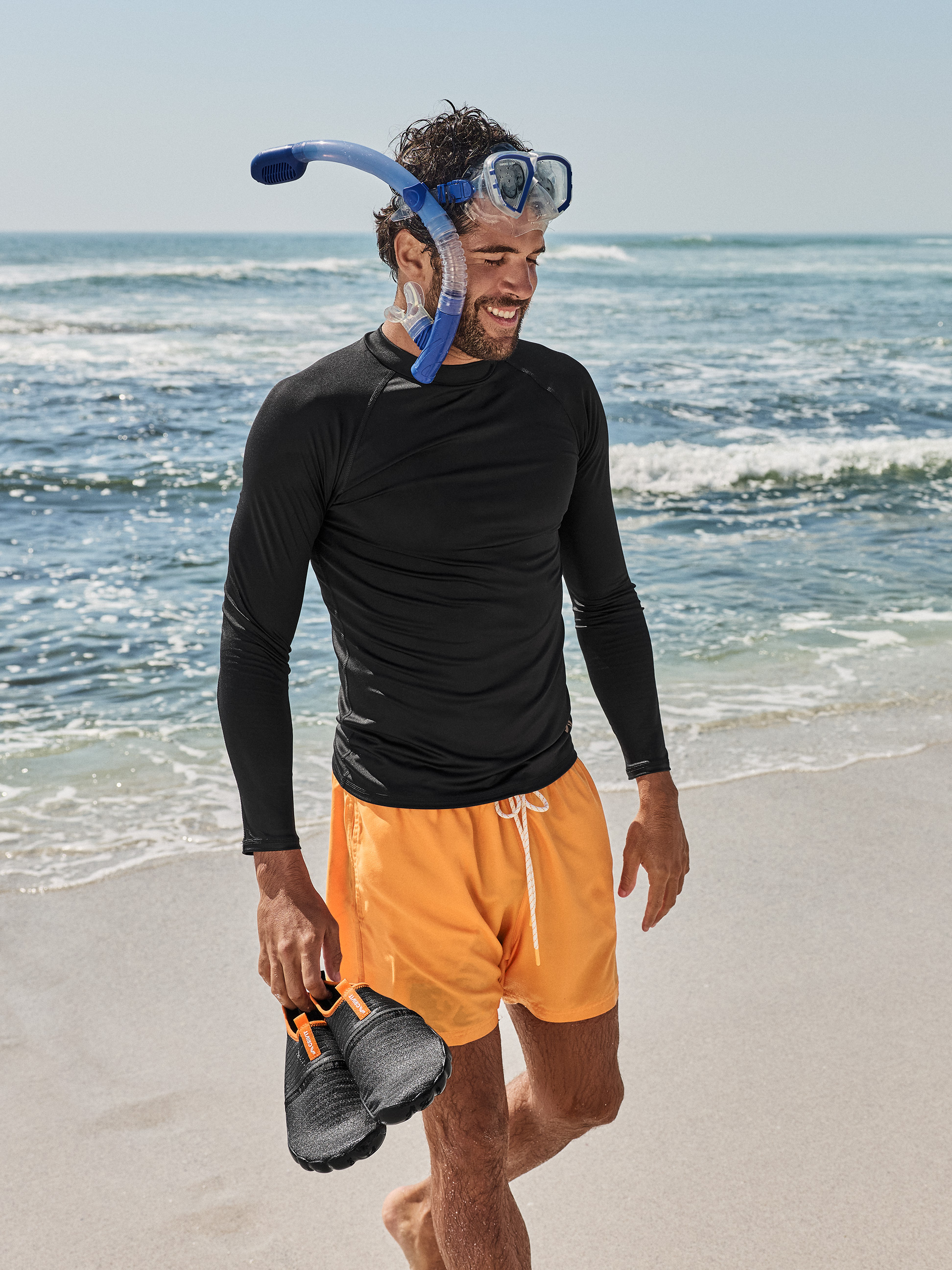 Mann in schwarzem Langarmshirt, orangefarbenen Badeshorts, mit Taucherbrille und Schnorchel, Wasserschuhe am Strand haltend.