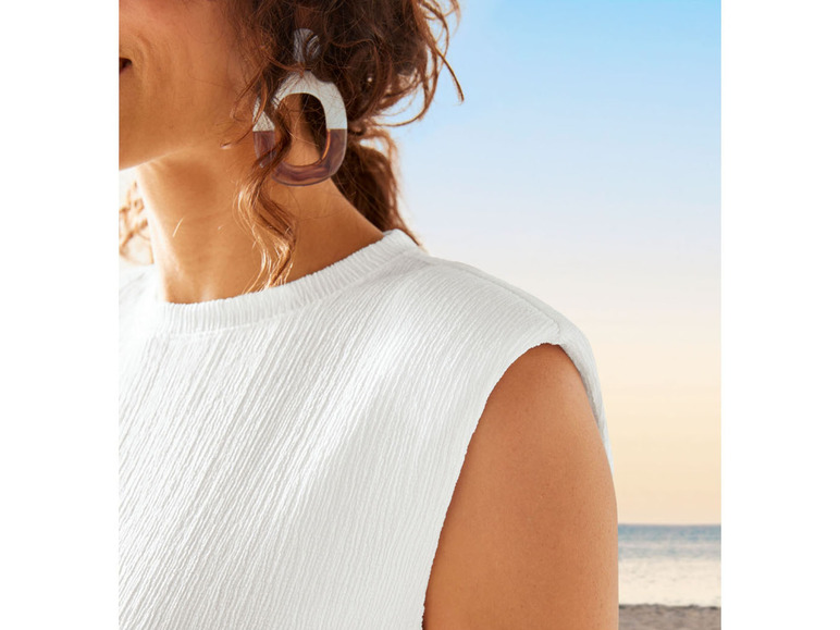 Eine Frau trägt ein weißes Oberteil mit Kapuze, vor dem Hintergrund eines Strandes und des Meeres.