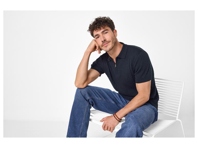 Mann in marineblauem Strick-Poloshirt und blauen Jeans sitzt auf einem weißen Stuhl.