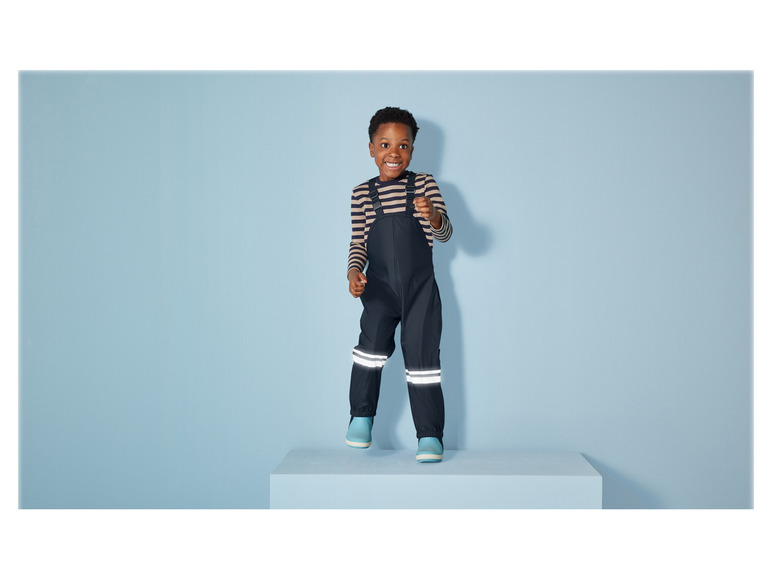 Ein Kind trägt eine schwarze Regenhose mit reflektierenden Streifen.