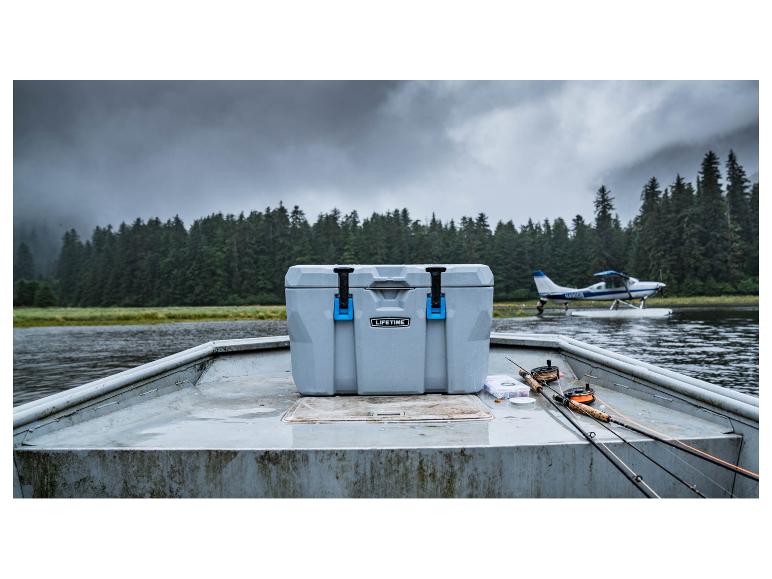 Graue Kühlbox auf einem Boot mit Angelruten, vor See- und Waldkulisse.