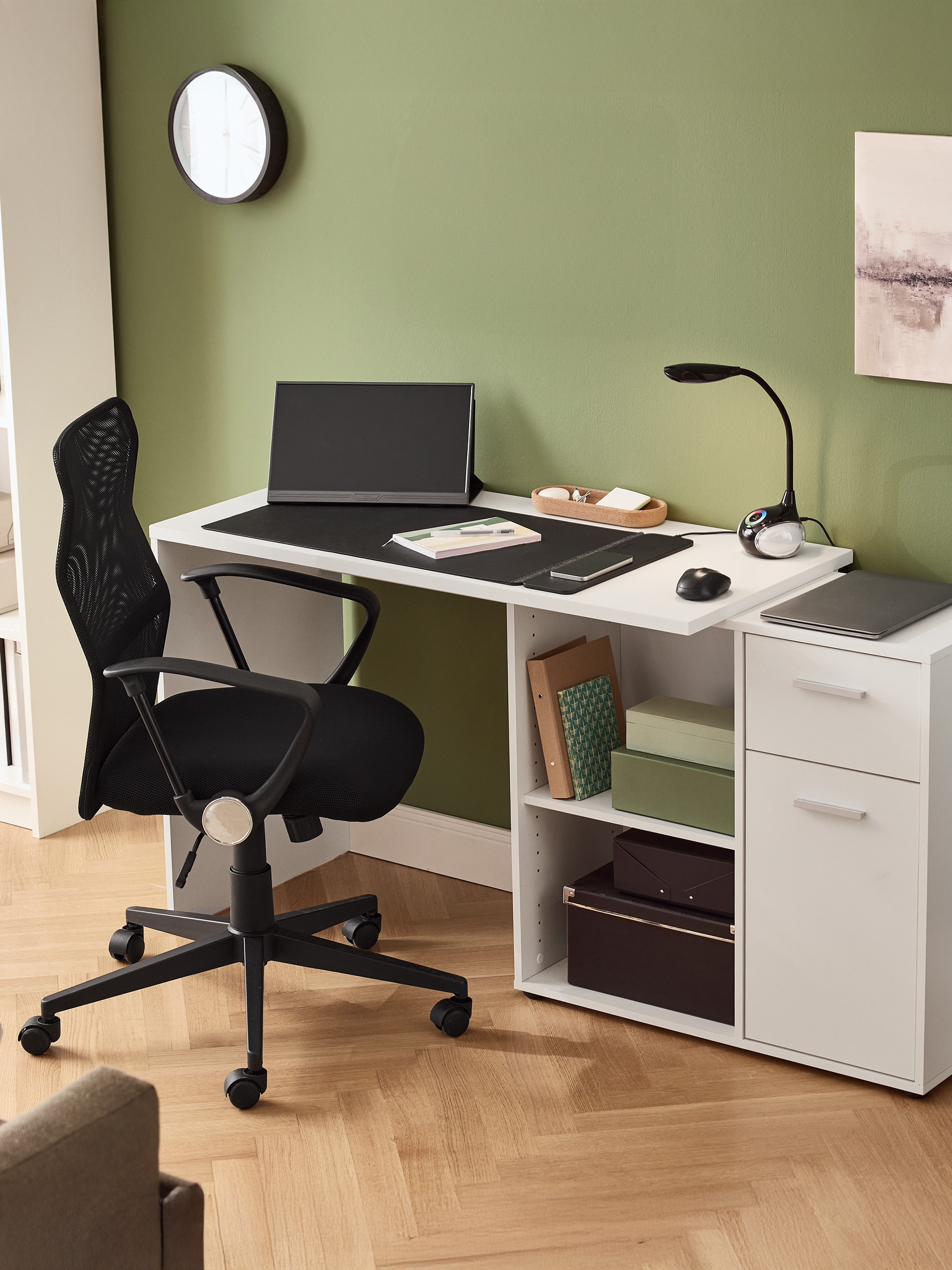Weißer Schreibtisch mit schwarzem Bürostuhl, Monitor, Lampe und Schubladen in einem grünen Raum.
