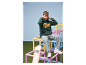 Junge in grünem 'Champs' Hoodie und ESMARA KIDS Jeans, sitzt auf einer bunten Struktur.