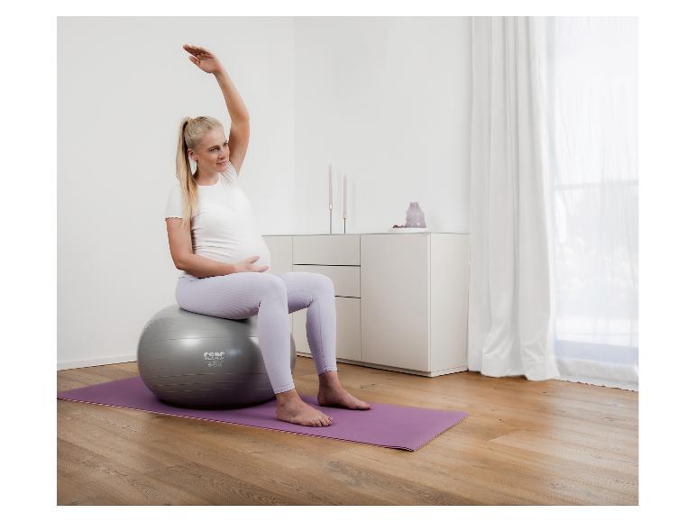 Schwangere Frau sitzt auf einem Gymnastikball auf einer Yogamatte und dehnt sich.