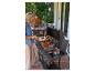Frau grillt Fleisch und Gemüse auf einem Gasgrill, im Hintergrund Familie am Tisch auf der Terrasse.