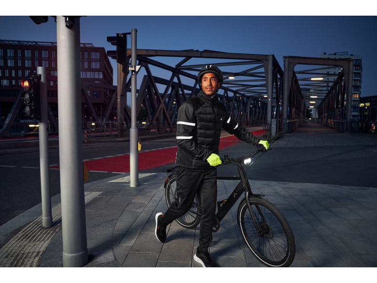Mann in reflektierender Fahrradjacke, Helm und Handschuhen schiebt ein Fahrrad in der Dämmerung.