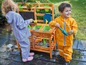 Zwei Kinder spielen in Regenkleidung im Garten mit einer Holzspielküche.