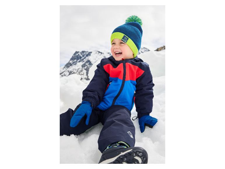 Glücklicher Junge in farbenfrohem Skianzug und Mütze sitzt im Schnee mit Bergen im Hintergrund.