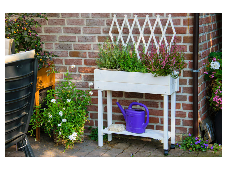 Ein weißes Pflanzenregal mit verschiedenen Blumen, einer Gießkanne und einem Spalier.