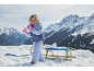 Mädchen in Skijacke, Hose und rosa Handschuhen, spielt mit Schnee neben einem Schlitten.