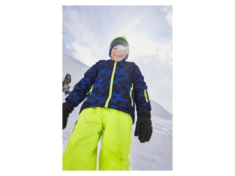 Lächelnder Junge in blauer Skijacke und neongelber Hose im Schnee.