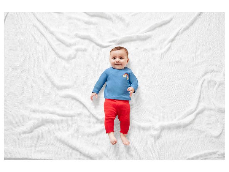 Ein Baby in einem blauen T-Shirt mit einem Regenbogen und roten Hosen liegt auf einer weißen Decke.