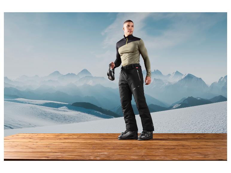 Ein Mann trägt eine Skihose und Skischuhe vor einer Berglandschaft.