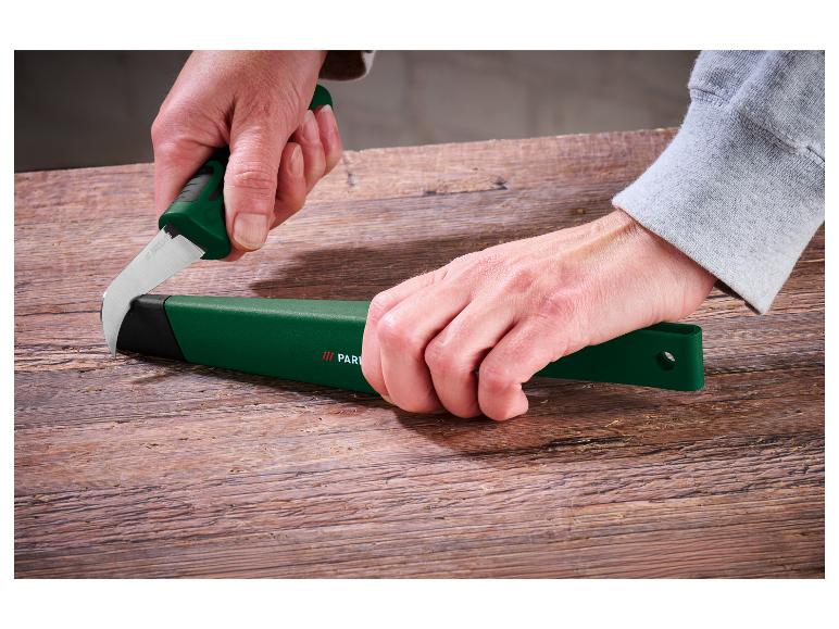 Hände stecken eine gebogene Klinge in eine grüne Scheide auf einem Holztisch.